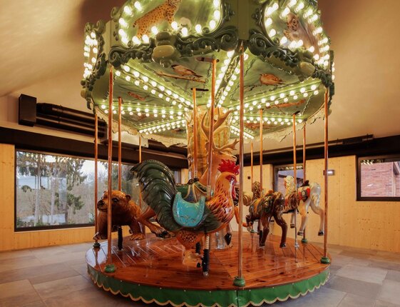 Warm-lit indoor vintage carousel with carved rooster, bear, horses and elephant on polished wooden platform in hotel family play area.