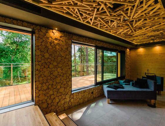 Cozy lounge with sofa, wooden lattice ceiling and log-slice accent wall opening to a sunlit forest-view terrace.