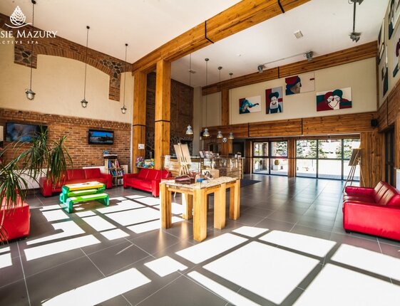 Sunlit spa hotel lobby with high wooden beams, exposed brick, red sofas, children's play table, central display table and glass doors