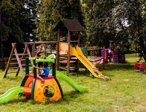 Family outdoor playground with wooden climbing frame, colorful slides and children playing on a grassy hotel garden.