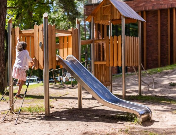 Outdoor children's playground: wooden playhouse, rope ladder and stainless slide, child climbing ladder among trees on hotel grounds.