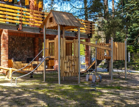 Outdoor wooden playground with slide, climbing nets and spring rocker next to hotel terrace with brick columns and trees
