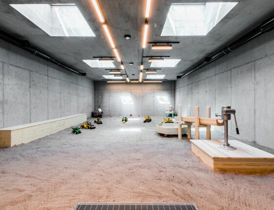 Indoor children's sand playroom with wooden play structures, toy tractors, skylights and concrete walls
