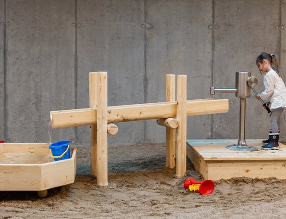 Indoor family play area with wooden water-play station and sandpit; child operating a hand pump, buckets and toys nearby.