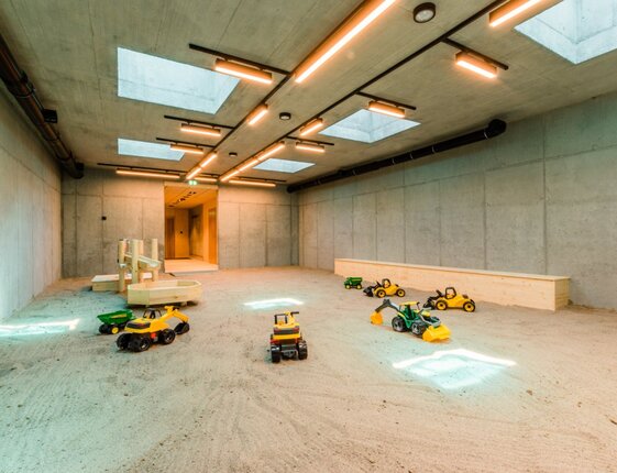 Family-friendly indoor sandbox with toy construction vehicles, wooden play structures, skylights and minimalist concrete walls.