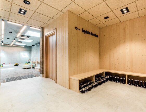 Wood-paneled boot room with bench, rows of black rubber boots and wall sign 'będzie brudno', opening to a skylit indoor playroom with toys.