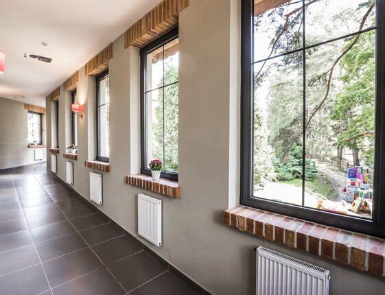 Bright hotel corridor with large windows, brick sills, tile floor, pink wall lamps and garden view with children's playground