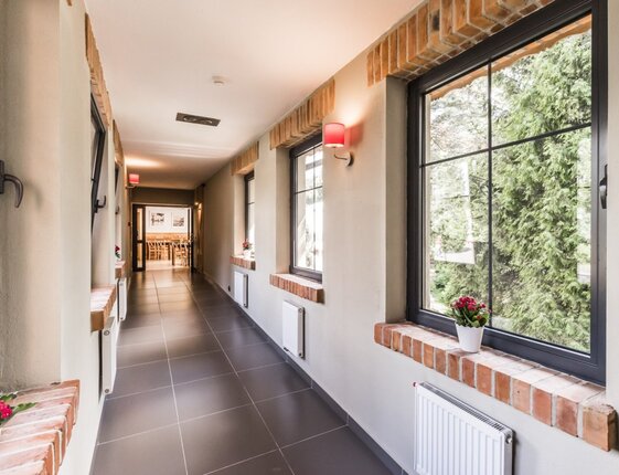 Bright hotel corridor with tiled floor, brick-framed windows, potted flowers, wall lamps and garden views toward dining area