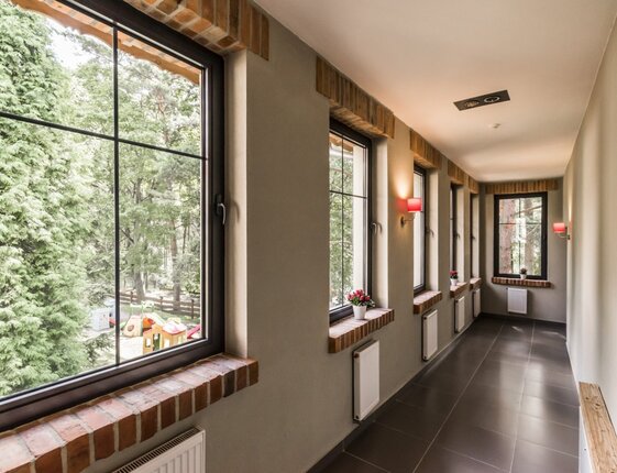Hotel corridor with large grid windows overlooking pine trees, brick sills with potted flowers, red wall sconces, radiators and tiled floor.