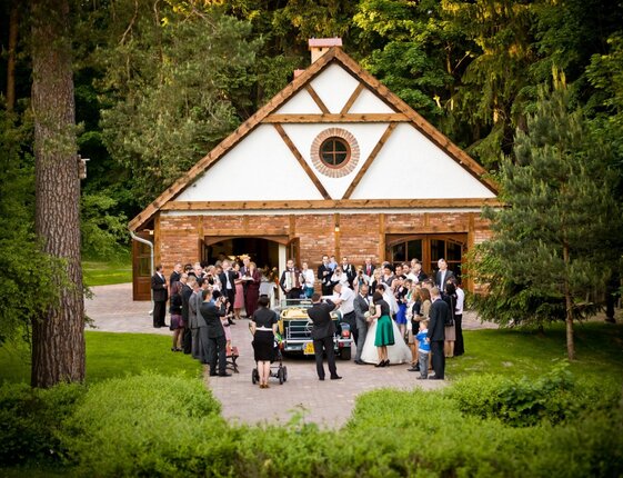 Rustic brick-and-wood wedding venue in forest clearing, guests gathered around a vintage car celebrating bride and groom on a manicured garden path.