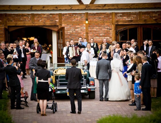 Bride and groom by vintage car outside brick reception hall, guests gathered, live accordion player and festive atmosphere