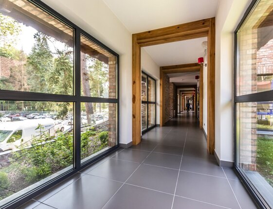 Bright hotel corridor with floor-to-ceiling windows, wooden frames and tiled floor, offering forest views and visible parking