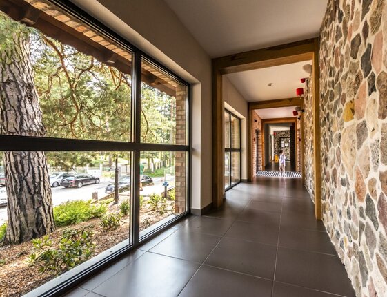 Sunlit hotel corridor with large windows facing pine trees and street, stone feature wall, tiled floor, child near end.