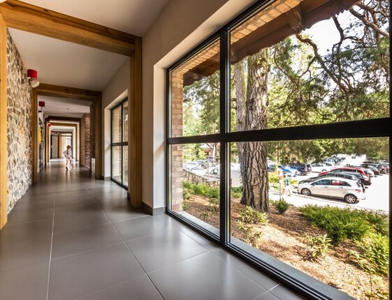 Bright hotel corridor with stone wall, wooden beams and floor-to-ceiling windows overlooking trees and parking; guest walking in distance.