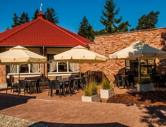Sunlit outdoor hotel terrace with cream umbrellas, black dining tables, brick facade and red-tiled roof among pines