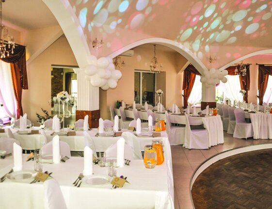 Elegant banquet hall for a wedding with white-draped tables, folded napkins, chandeliers, balloon columns, colored lights and curved dance floor