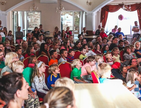 Packed hotel event hall with families and children seated on red chairs, watching a lively kids' show under chandeliers and arched ceiling.