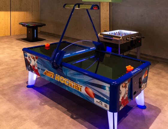 Modern game room with illuminated air hockey table, foosball table and arcade station on polished floor.