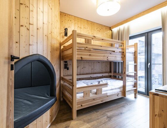 Hotel room with pine bunk beds, wood-paneled walls, built-in black leather reading nook, large window and balcony.