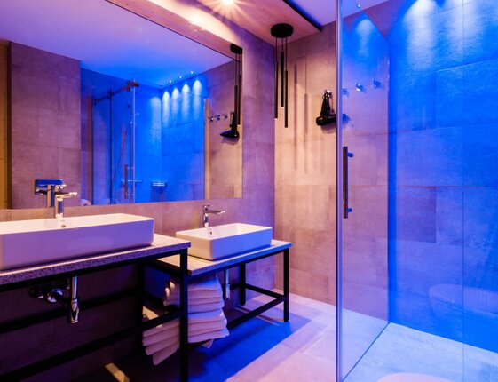 Modern hotel bathroom with dual rectangular sinks, large mirror, glass walk-in shower, stacked towels and blue mood lighting.