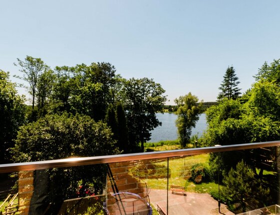 Hotel balcony with glass railing overlooking a tranquil lake, leafy trees and sunlit park benches