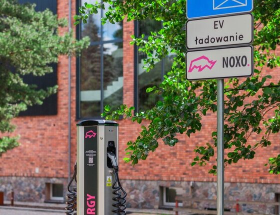 Outdoor electric vehicle charging station branded NOXO ENERGY beside hotel parking, brick façade and greenery