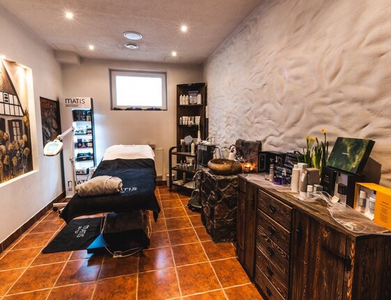 Cozy spa treatment room with heated massage table, soft lighting, stone basin, skincare products and rustic wooden cabinetry.