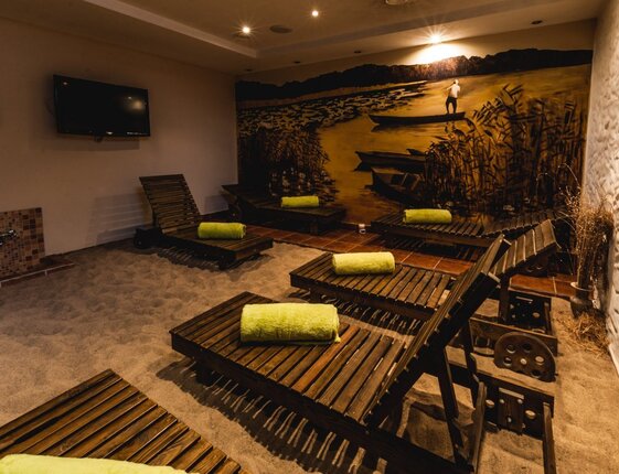 Spa relaxation room with wooden lounge chairs on sand-like floor, lime towels, warm mural of boats and soft ambient lighting