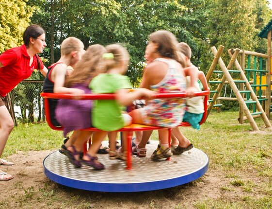 Outdoor hotel playground with spinning roundabout full of children, staff member pushing, wooden climbing frame and slide nearby.