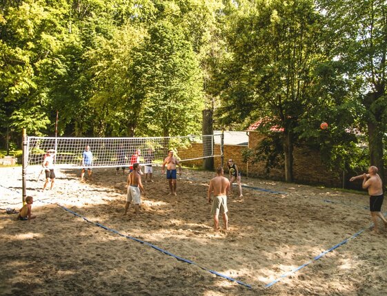 Guests playing beach volleyball on a shaded sand court surrounded by trees and resort grounds