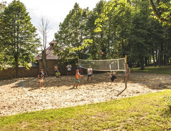 Guests playing casual beach volleyball on a shaded sand court amid trees and grassy resort grounds.