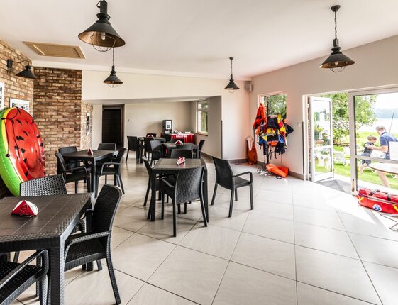 Lakeside activity room with black wicker tables, brick wall, pendant lamps and life jackets; open door shows a parent carrying a child.