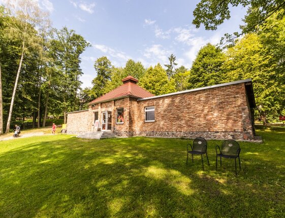 Single-story brick building with red-tiled roof on a grassy lawn, two outdoor chairs; guests stroll past with a stroller