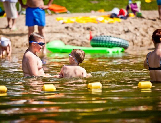 Children and adults wading at a sandy lakeside beach, two boys in goggles in shallow water, inflatable floats and yellow buoys