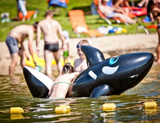 Family-friendly lakeside beach with children playing on a black inflatable orca, sunloungers and swimmers in background.