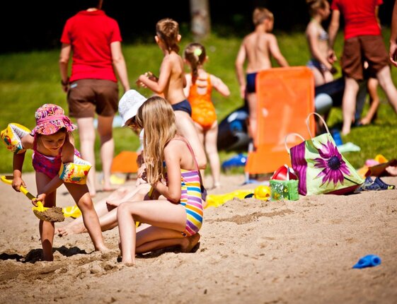 Family-friendly sandy beach with children digging in sand, colorful lounge chairs, inflatable armbands and a floral beach bag nearby.