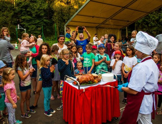 Chef presenting roast pig on a red cart to excited children and families at an outdoor hotel event under a canopy