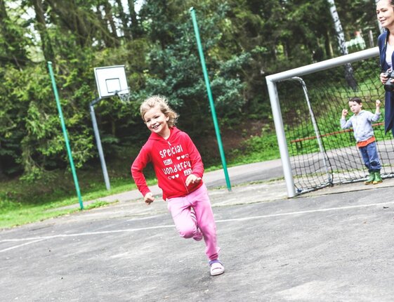 Smiling girl in red hoodie runs on outdoor sports court by a goal and hoop; woman with camera and a boy watch.