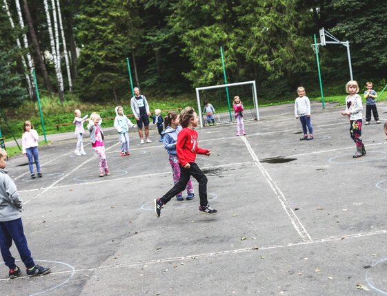 Children playing on an outdoor sports court marked with chalk circles, a supervisor nearby and forested backdrop — family-friendly group activity.