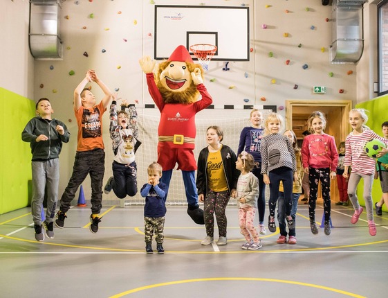 Children playing with a hotel mascot in a bright indoor activity hall with climbing wall, basketball hoop and padded walls.
