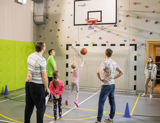 Family activity hall with kids shooting a basketball under a hoop, supervised staff, climbing-wall holds and padded green walls.