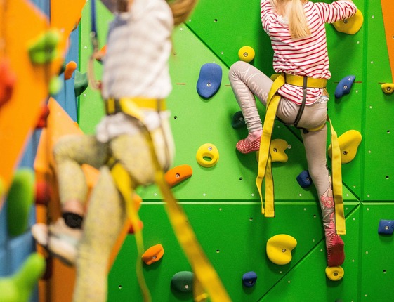 Two children in harnesses climbing a bright green indoor rock wall with colorful holds in a family play area.