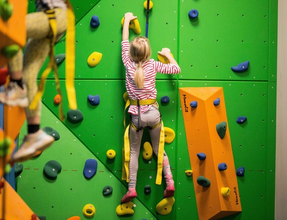 Children's indoor climbing wall with bright green panels, colorful holds and safety harness; child in striped shirt climbing in family activity area.