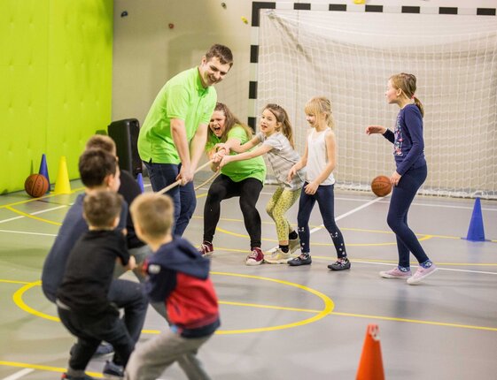 Children and two staff play tug-of-war in a bright indoor sports hall with goal net, cones and basketballs.