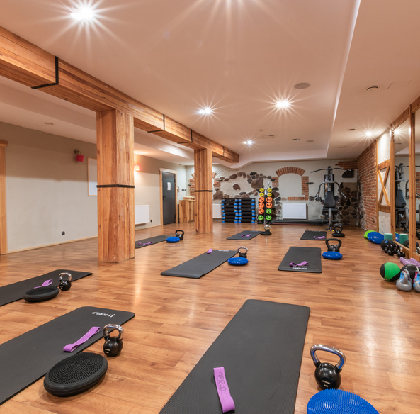 Przytulne hotelowe studio fitness z drewnianymi belkami, matami do jogi, kettlebellami, poduszkami równoważnymi, lustrem i ceglaną ścianą.