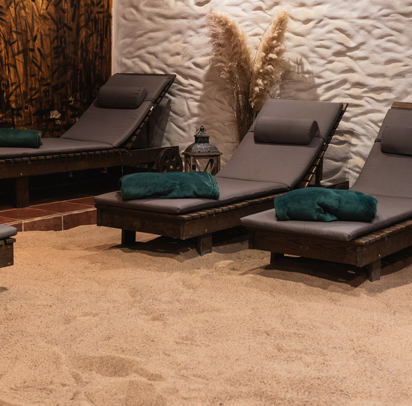 Strefa relaksu spa z drewnianymi leżankami na piaszczystej podłodze, szare poduszki, zielone ręczniki i ozdobne trawy