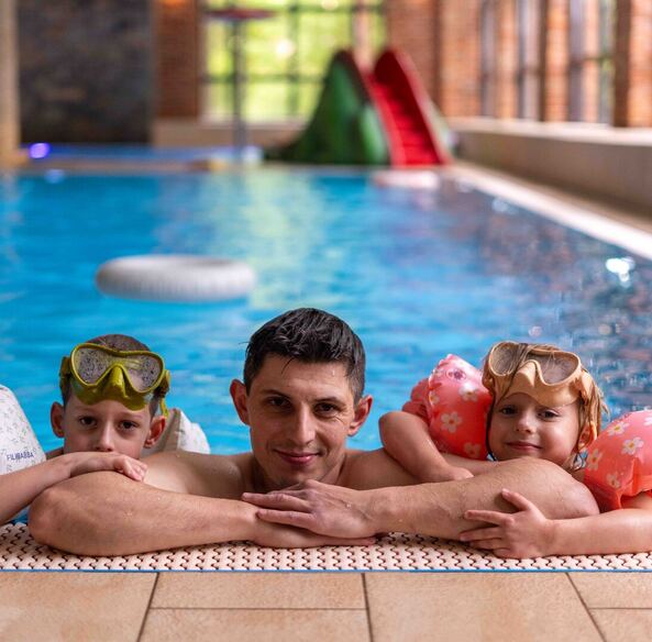 Father with two children at edge of indoor hotel pool, kids in floaties and goggles, slide and large windows behind.