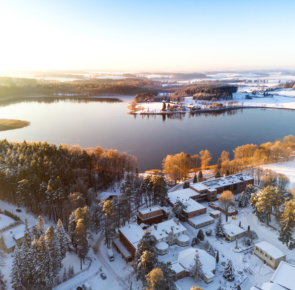 Lakeside hotel and resort buildings nestled in a snowy pine forest beside a calm winter lake at sunrise.