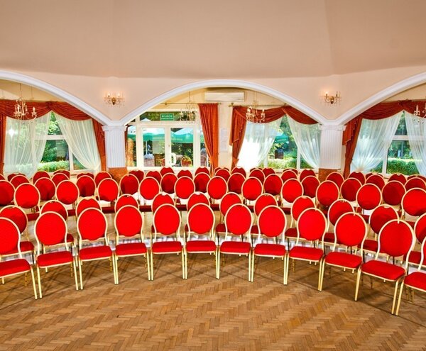 Elegancka sala bankietowa hotelu z rzędami czerwonych krzeseł, łukowymi oknami, żyrandolami, parkietem i widokiem na ogród.