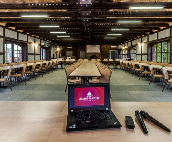 Przestronna, rustykalna sala konferencyjna z długimi stołami, drewnianymi belkami, kominkiem i laptopem z mikrofonami